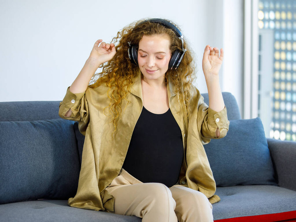 Coiffure bouclée jeune heureuse femme enceinte caucasienne en tenue décontractée et casque assis sur un canapé confortable en écoutant des chansons de musique en streaming en ligne. - Photo, image