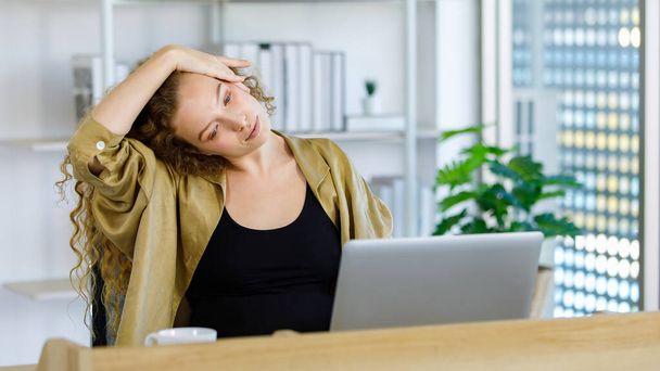 Curly hairstyle young happy healthy Caucasian pregnancy mother in casual outfit sitting at working desk with laptop computer and coffee cup at workspace stretching relaxing resting arms and hands. - 写真・画像