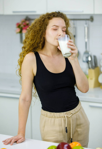 Feliz saludable peinado rizado joven Caucásico embarazo madre modelo en traje casual de pie sosteniendo vaso de leche en las manos para el desayuno en la cocina completamente amueblada en casa - Foto, imagen