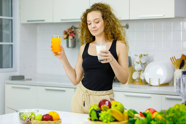 Glückliche gesunde lockige Frisur junge kaukasische Schwangerschaftsmutter Modell in lässigem Outfit stehend mit einem Glas Orangensaft und Milch in der Hand zum Frühstück in der voll ausgestatteten Küche zu Hause. - Foto, Bild