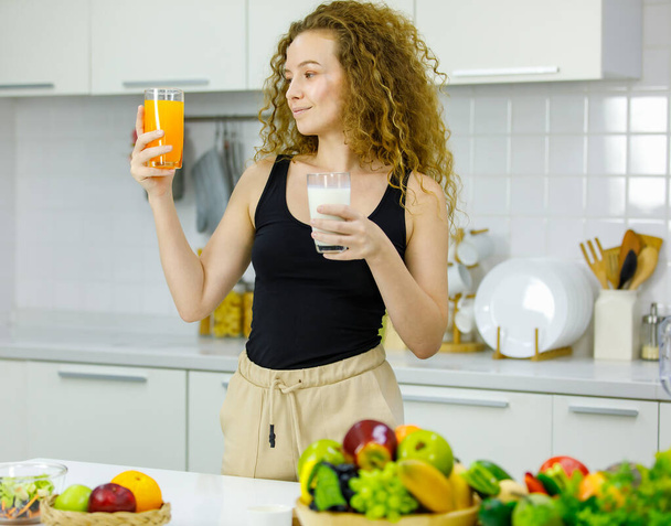 Happy healthy curly hairstyle young Caucasian pregnancy mother model in casual outfit standing holding choosing glass of orange juice and milk in hands for breakfast in full furnished kitchen at home. - Photo, Image
