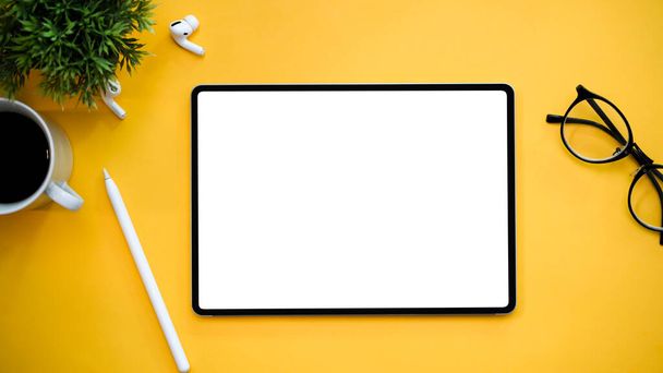 Flache, moderne Arbeitsfläche mit Tablet-Display-Attrappe, Brille, Stift, Ohrhörer, Kaffeetasse und Dekorationsanlage auf gelbem Hintergrund. Ansicht von oben - Foto, Bild