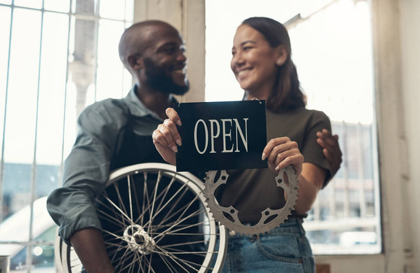 Tu moto está en buenas manos. Foto de una joven pareja de pie juntos en su tienda de bicicletas y sosteniendo un cartel abierto. - Foto, imagen