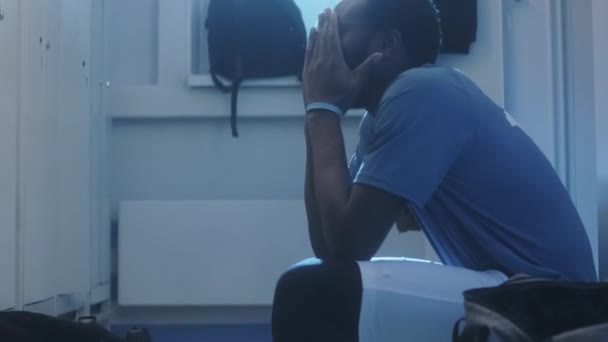 Widok z boku przechyla się młody czarny mężczyzna sportowiec siedzi przy szafkach w szatni, przygotowuje się psychicznie, jest zdenerwowany przed meczem wcześnie rano - Materiał filmowy, wideo