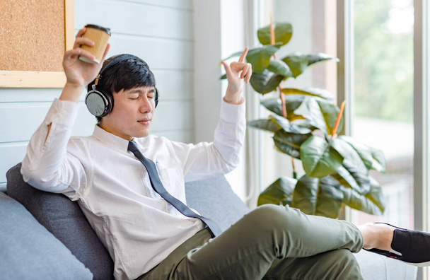 Asiatische junge hübsche professionelle erfolgreiche männliche Geschäftsmann Mitarbeiter in formalen Business-Hemd und Krawatte sitzt auf einem gemütlichen Sofa und hört Streaming-Musik im Internet surfen mit dem Smartphone. - Foto, Bild