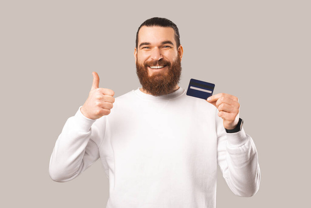 Homem barbudo extático está mostrando o polegar para cima enquanto segura um cartão de crédito. Estúdio filmado sobre fundo branco. - Foto, Imagem