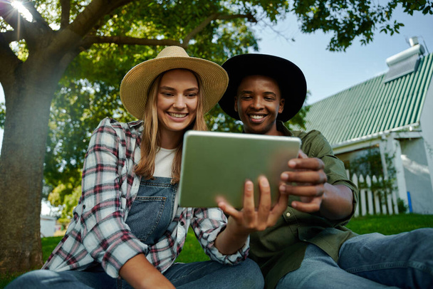 Mixed race male and female famers sitting together on green grass looking at digital tablet - 写真・画像