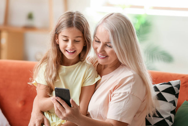 Little girl with her grandma using mobile phone on sofa at home - Фото, изображение