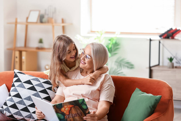 Маленькая девочка со своей бабушкой читает книгу на диване дома - Фото, изображение