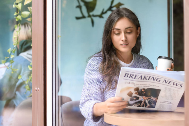 Beautiful young woman with cup of coffee reading newspaper at table in cafe - Photo, Image