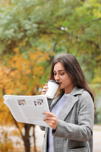 Beautiful young woman drinking coffee and reading newspaper in autumn park - Фото, изображение