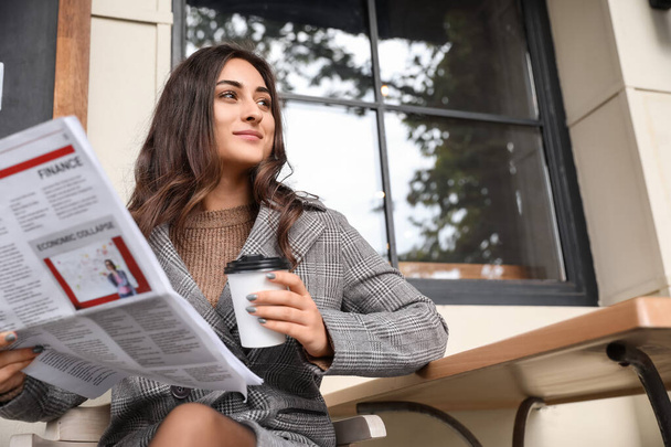 Gyönyörű fiatal nő csésze kávéval és újságpapírral ül az utcai kávézóban - Fotó, kép