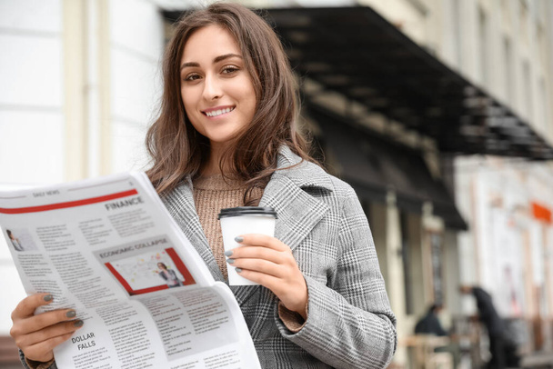 Beautiful young woman with cup of coffee and newspaper on street - Photo, Image