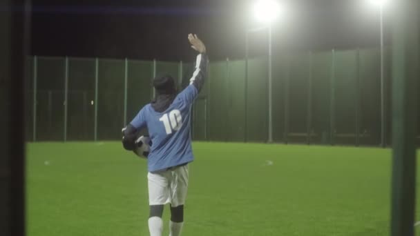 Giovane uomo nero indossa abbigliamento sportivo e cappello caldo, tenendo la palla, entrando campo da calcio vuoto, salutando pubblico invisibile, sognando di essere giocatore famoso - Filmati, video