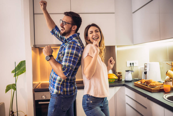 Νεαρό ζευγάρι στη σύγχρονη κουζίνα, χορεύοντας και γελώντας, ευτυχισμένη νεαρή γυναίκα και ο σύζυγός κινείται προς την αγαπημένη μουσική. - Φωτογραφία, εικόνα