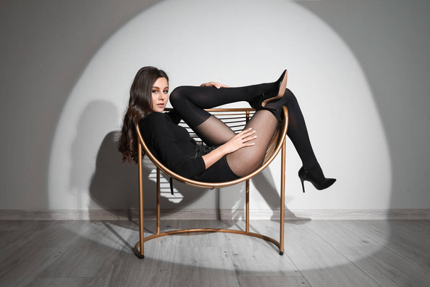 Gri duvarın yanındaki koltukta oturan siyah taytlı seksi genç kadın. - Fotoğraf, Görsel