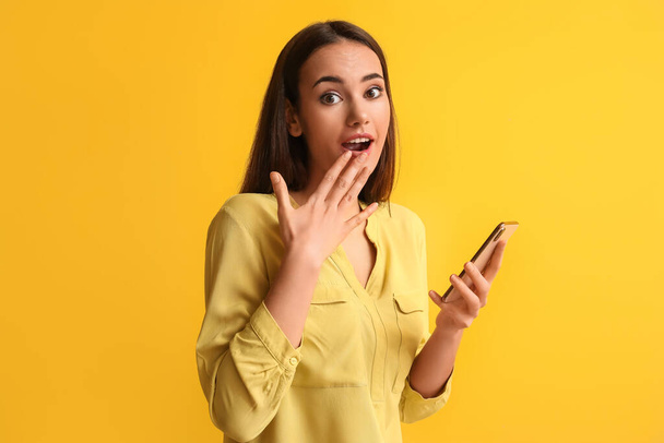 Beautiful surprised young woman with mobile phone on yellow background - Photo, image