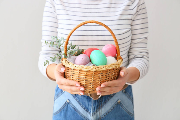 Woman holding gift basket with painted Easter eggs on light background, closeup - Foto, imagen