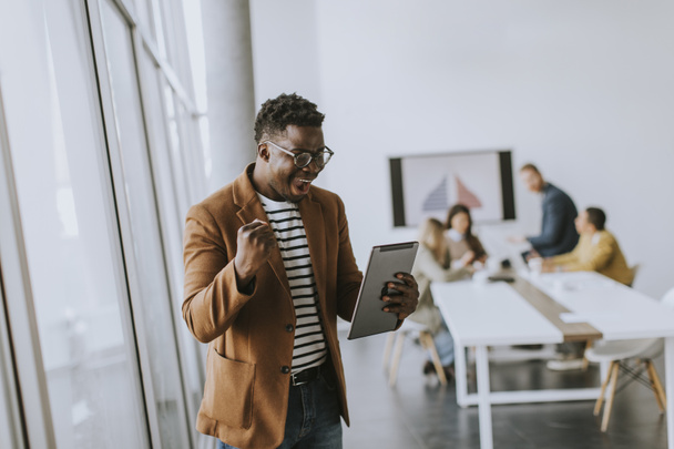 Schöner junger afroamerikanischer Geschäftsmann, der mit einem digitalen Tablet arbeitet und vor seinen Mitarbeitern im Sitzungssaal gute Nachrichten erhält - Foto, Bild