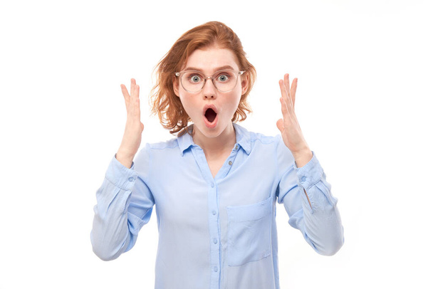 Shocked face red-haired girl in business shirt looks surprised with open mouth spreads hands on white studio background - Foto, Bild