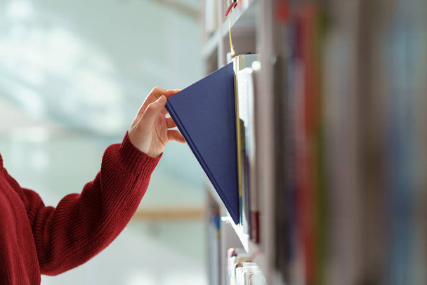Woman hand picking book from bookshelf in library in university, college, high school or bookshop - Photo, Image