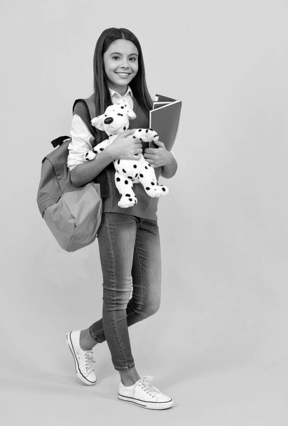 щаслива дівчина школи-підлітка з рюкзаком та копіювальною книгою тримає іграшку на жовтому фоні, урок
 - Фото, зображення