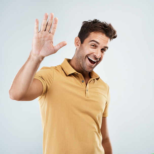 Are we going to do a high five. Portrait of a cheerful young man smiling brightly with his one hand in the air while standing against a grey background. - Foto, imagen