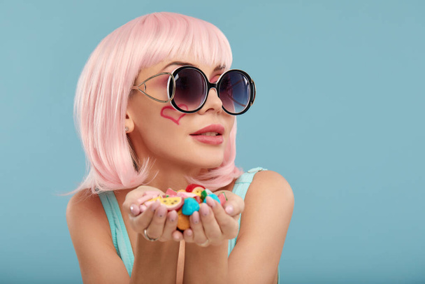 Close up of a flirtatious female model on blue background wearing a pink wig holding different kinds of candies. Pretty glamorous woman pink hair charm sweets lifestyle enjoyment sugar - 写真・画像