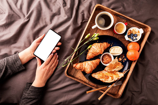 Woman using smartphone while eating continental breakfast in bed. Wooden tray with coffee and croissants on grey sheets from above (top view, flat lay). Blank screen copy space. - Foto, imagen