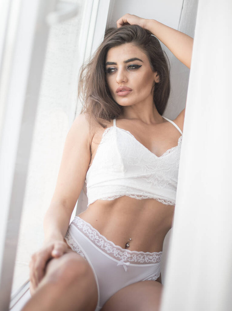 Attractive sexy brunette in white lingerie  posing provocatively, near a window,studio shot. Portrait of sensual woman with long hair and white bikini in classic boudoir scene. - Foto, afbeelding