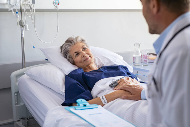Doctor assuring senior woman patient lying on hospital bed. Happy elderly patient lying on hospital bed feeling relaxed after doctor giving health update. Physician examining hospitalized mature woman lying in hospital room before surgery while holdi - Фото, изображение