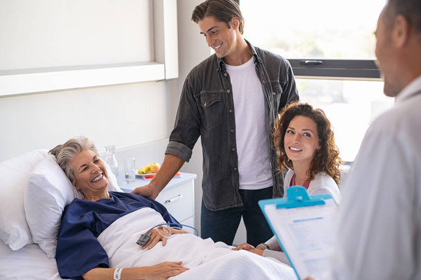Doctor holding clipboard talking to family and smiling patient. Happy senior woman lying on hospital bed with lovely son and daughter visiting and talking to doctor. Professional friendly physician gives the results of the medical report to patient's - Fotoğraf, Görsel