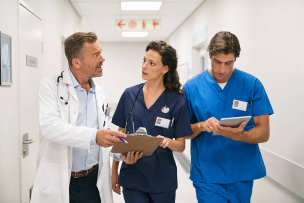 Medical team discussing medical report on digital tablet while walking in a corridor of modern hospital. Mature doctor and surgeon working on digital tablet with nurse and walking in hospital hallway. Head physician working with his medical staff at  - Foto, Imagem