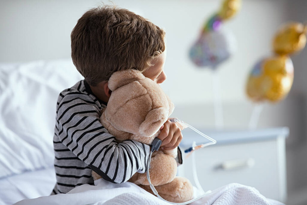 Rear view of scared little boy with intravenous drip in hand hugging teddy bear sitting on hospital bed. Sick lonely child looking through the window in clinic pediatric ward while embracing his favorite toy with pulse oximeter on finger. Unwell sad  - Zdjęcie, obraz
