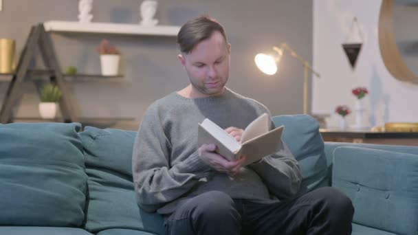 Casual Man Czytanie książki podczas siedzenia w kanapie - Materiał filmowy, wideo