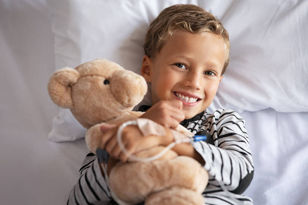 Happy little boy hugging teddy bear with drip in hand smiling on hospital bed. Portrait of smiling child embracing teddy bear on gurney at private clinic. Cute kid with stuff toy lying on hospital bed and looking at camera with big grin. - Фото, зображення