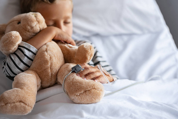 Little cute boy hugging teddy bear with intravenous drip in hand, sleeping under bed sheet in hospital. Sick child with pulse oximeter on finger resting with his toy on gurney at pediatric ward with copy space. Lovely kid embracing stuff toy lying on - Photo, Image