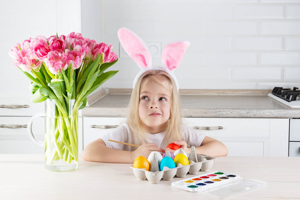 Una chica de ensueño con orejas de conejo pinta huevos de Pascua en una mesa de madera. - Foto, Imagen