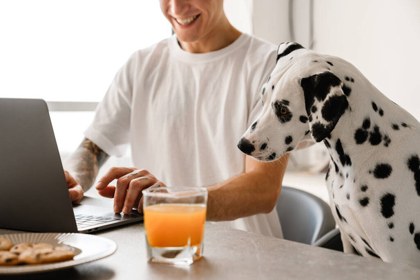 Smiling mid aged man working on laptop computer while sitting at the kitchen wit his dog, having breakfast - 写真・画像
