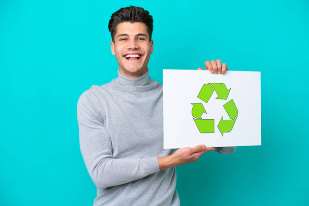 Junger gutaussehender kaukasischer Mann isoliert auf blauem Grund mit einem Plakat mit Recycling-Ikone und glücklichem Gesichtsausdruck - Foto, Bild