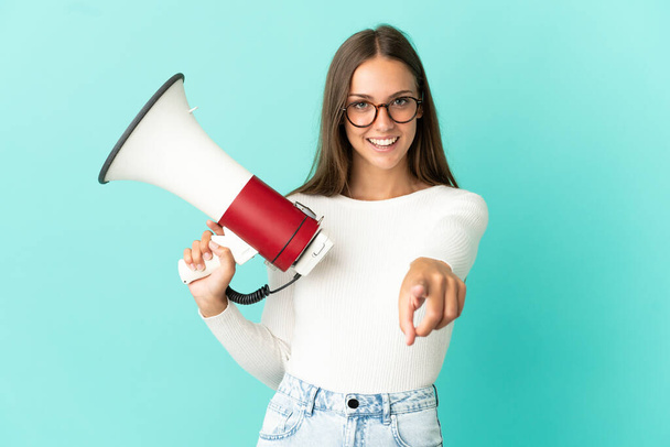 Young woman over isolated blue background holding a megaphone and smiling while pointing to the front - Foto, Bild