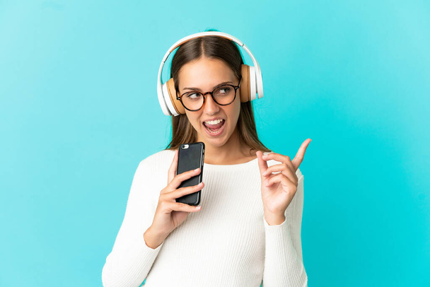 Fiatal nő elszigetelt kék háttér hallgat zenét egy mobil és énekel - Fotó, kép