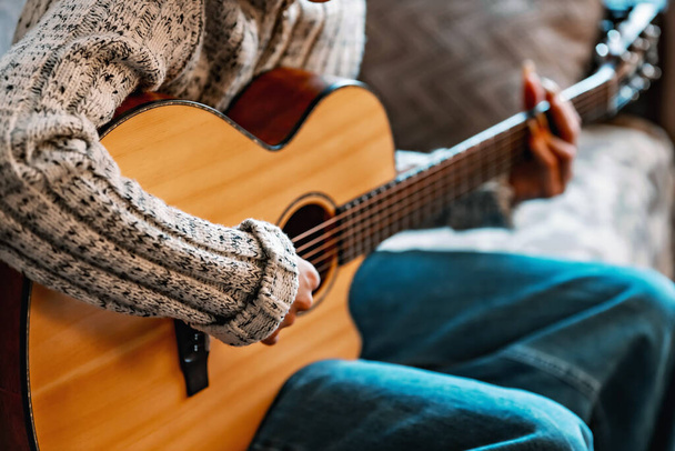 Молодая девушка с длинными ногтями играет на акустической гитаре дома. Подросток сидит на диване в комнате и учится играть на гитаре - Фото, изображение