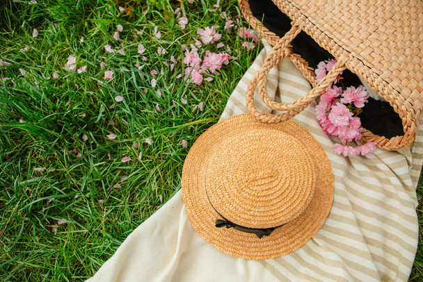 Picknickdecke mit Strohhut und Tasche auf grünem Gras bedeckt mit rosa Sakura-Blumen - Foto, Bild