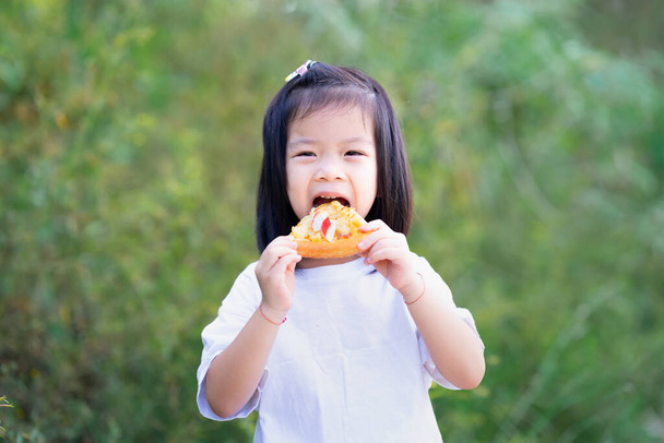 Portré aranyos ázsiai lány élvezi eszik pizza gusto. Boldog gyerek, aki ételt tart a kezében, harapással a szájába veszi. A gyerekek fehér pólót hordanak. Zöld természet háttér. - Fotó, kép