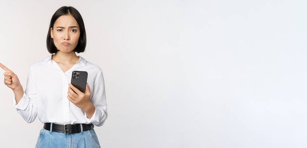 Skeptisch Aziatisch modern meisje, met smartphone, wijzende vinger links naar lege ruimte en fronsend aarzelend, heeft twijfels, staande over witte achtergrond - Foto, afbeelding