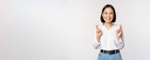 Retrato de mujer joven coreana, chica asiática cruzan los dedos y rezan, pidiendo deseo, anticipando, esperando resultados, de pie sobre fondo blanco - Foto, Imagen