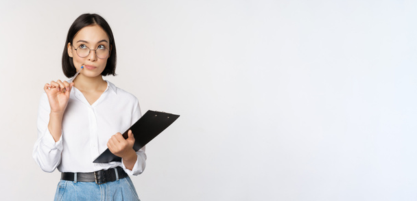 Asian girl in glasses thinks, holds pen and clipboard, writing down, making notes, standing over white background - Photo, image