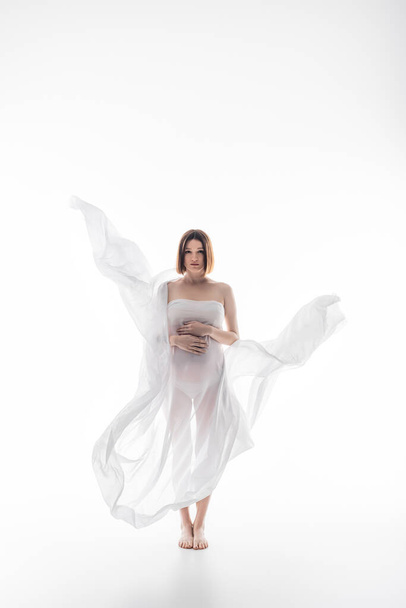 Full length of pregnant woman in fabric touching belly on grey background - Foto, immagini