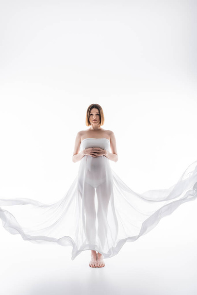 Έγκυος γυναίκα σε ύφασμα που αγγίζει την κοιλιά και ποζάρει σε γκρι φόντο - Φωτογραφία, εικόνα